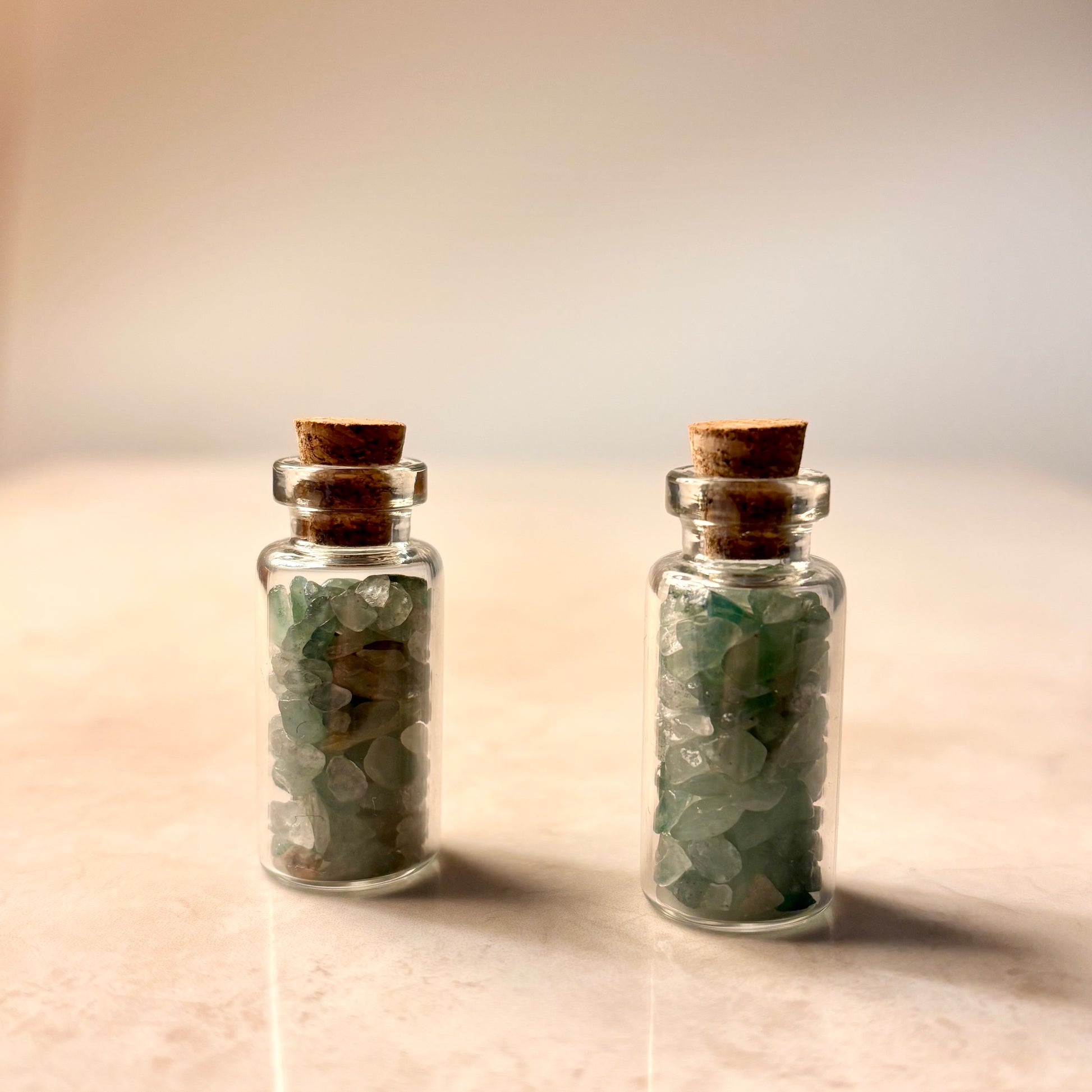 Two small glass bottles with cork lids containing green stones on a beige background