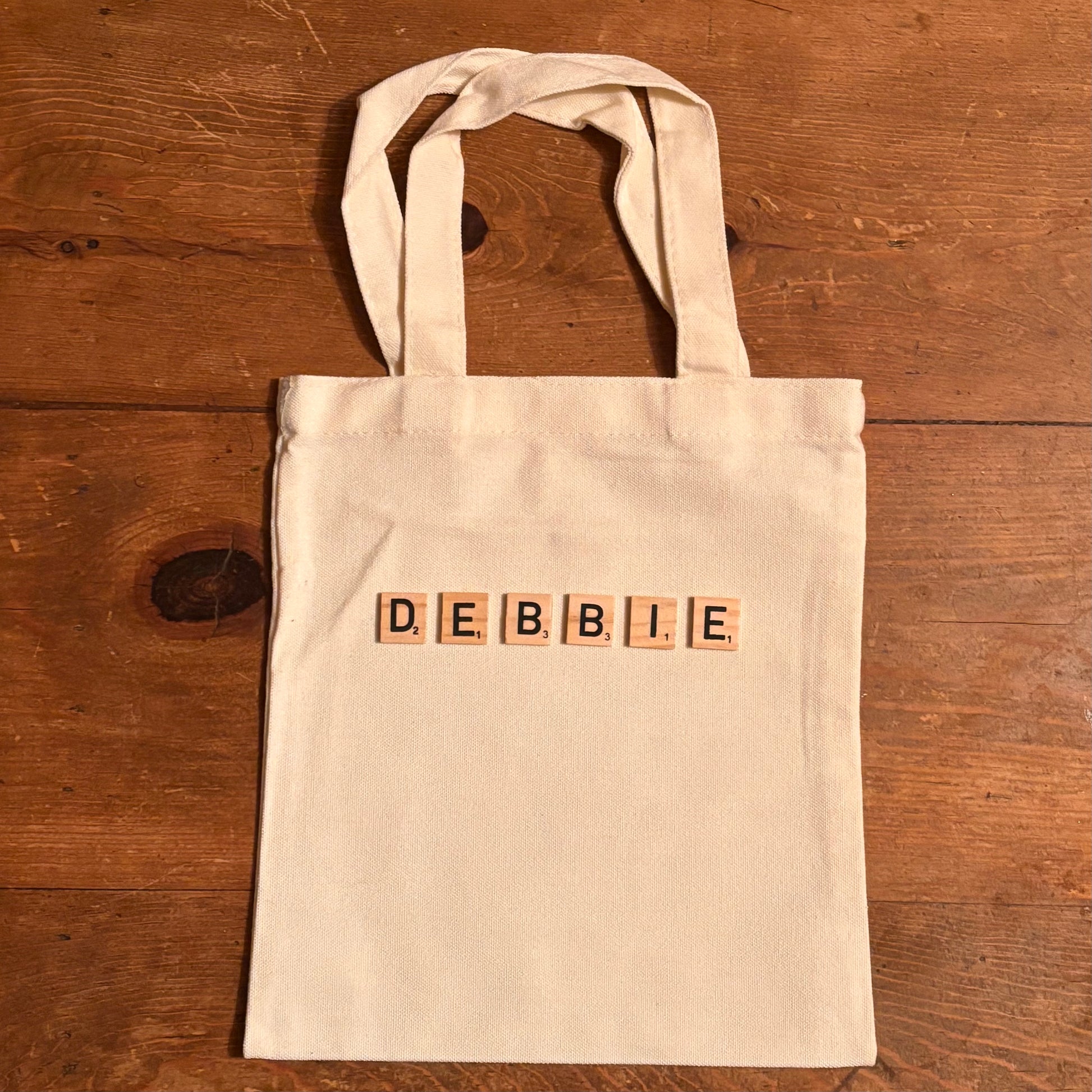 Beige tote bag with 'DEBBIE' spelled out using wooden blocks on a wooden surface
