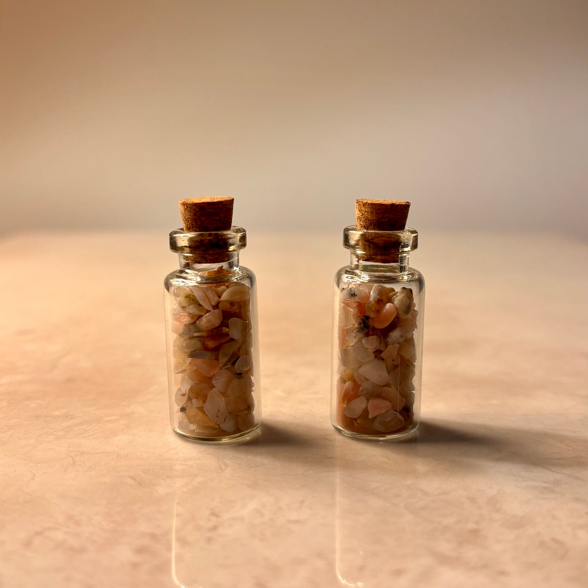 Two small glass jars with cork lids containing small pink stones on a beige surface