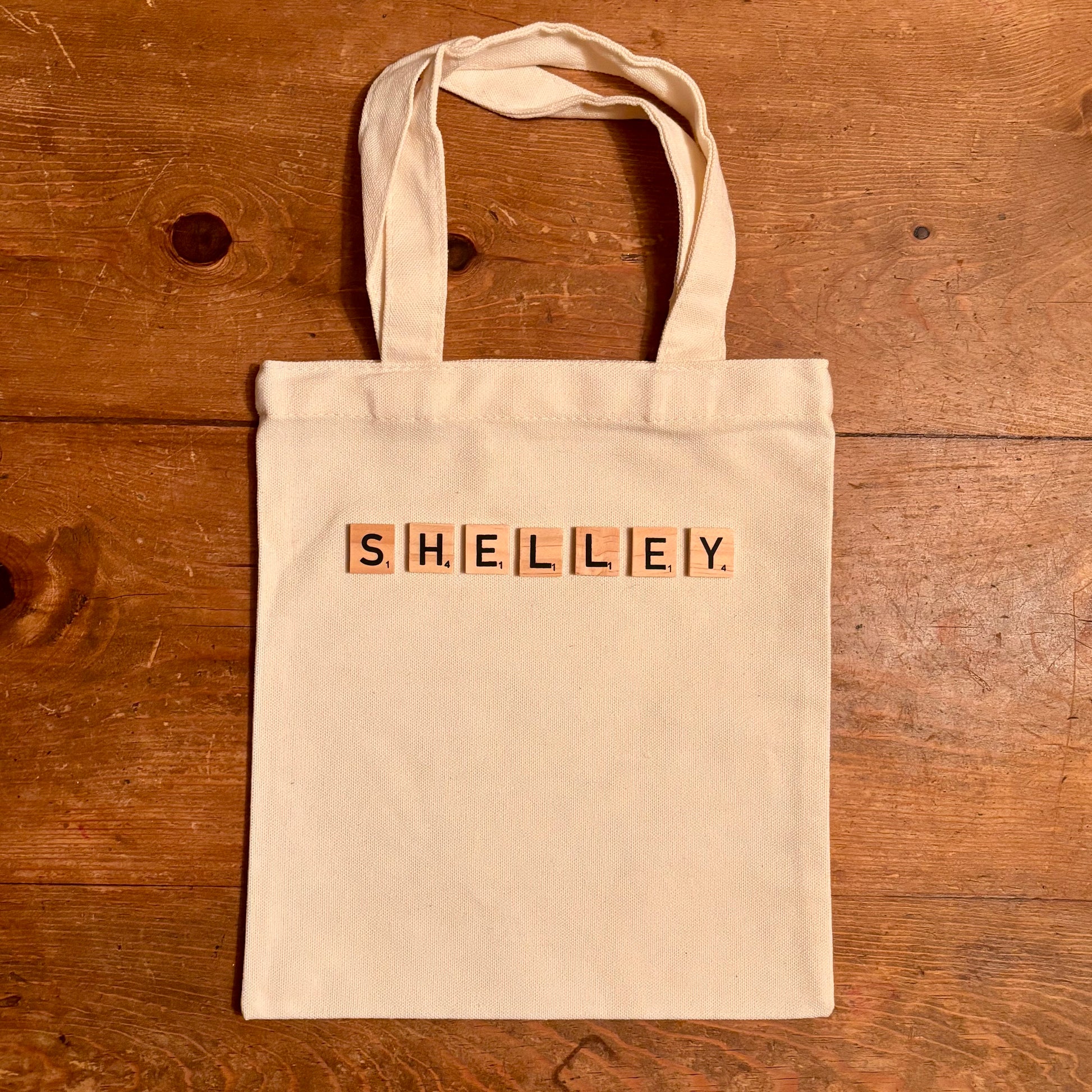 Beige tote bag with 'SHELLEY' spelled out using wooden blocks on a wooden surface