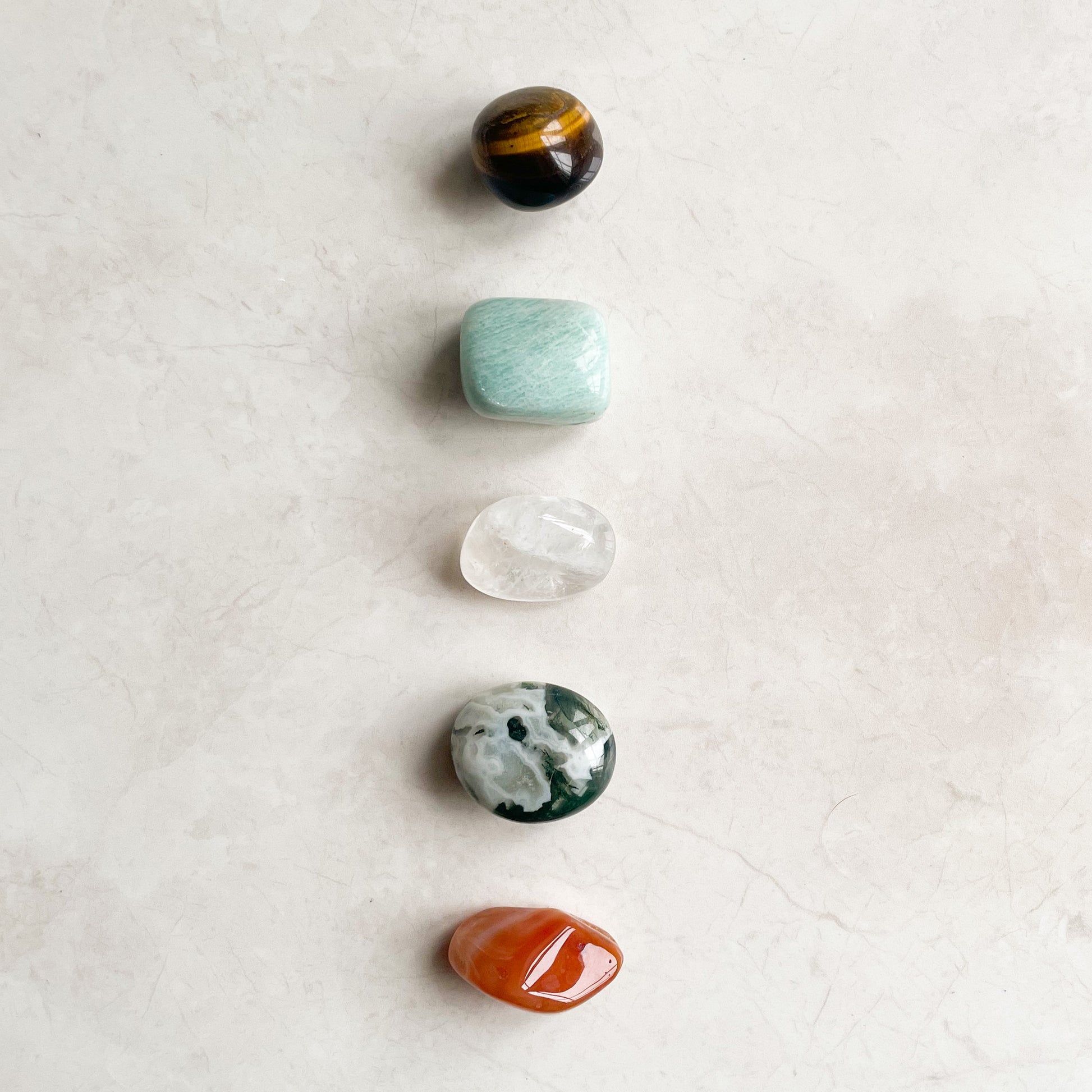 An arrangement of five tumbled crystals on a light surface, from top to bottom: black with hints of orange, translucent white, green with white patterns, clear with inclusions, and orange.