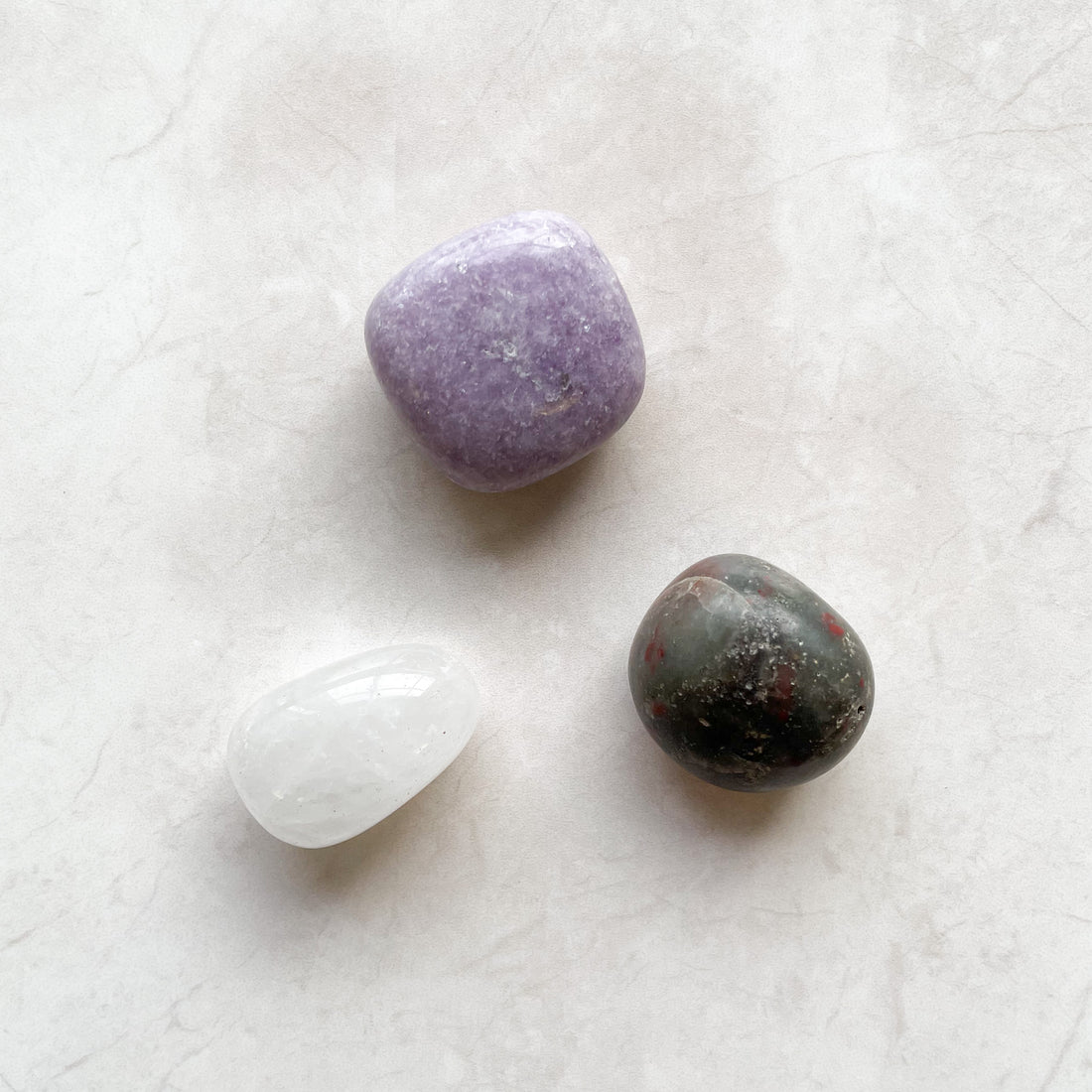 three crystal tumbles, lepidolitte which is a pastel purple, clear quartz which is white and bloodtone which is grey, red with speckles of colour