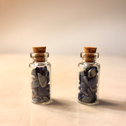 Two small glass jars with cork lids containing bliue lapis crystal chips stones on a gradient background
