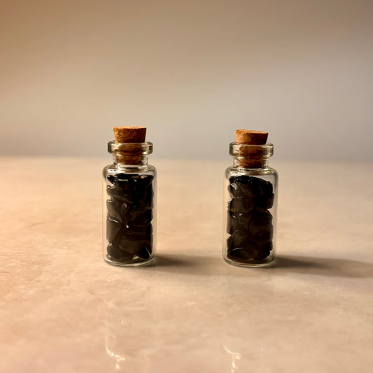 Two small glass bottles with cork lids containing dark black crystal chips on a beige surface.