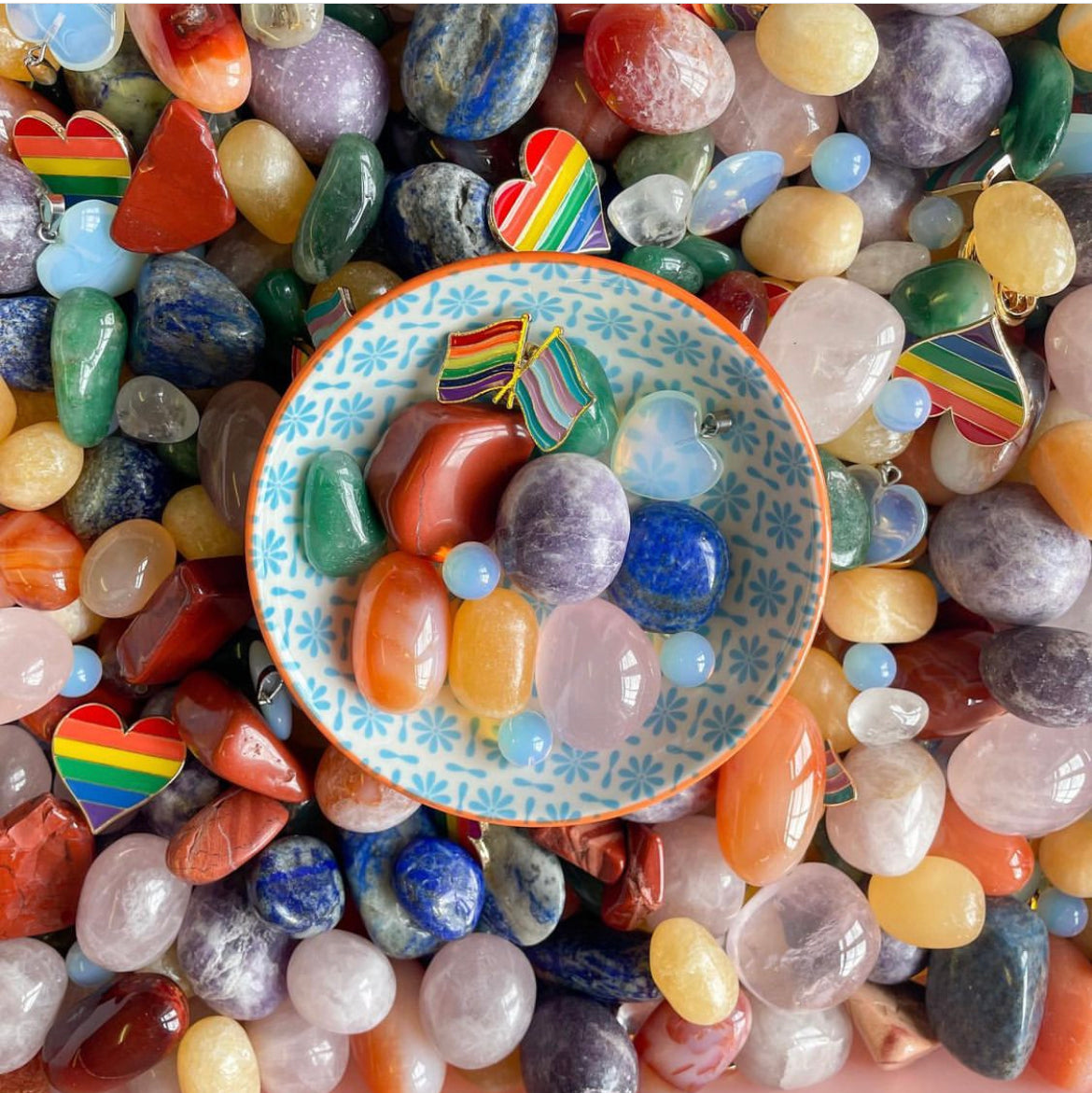 a collection of rainbow crystals for pride month with red jasper, organge calcite, green aventurine, lapis lazuli, lepidolite, rose qaurtz and opalite with a rainbow flag badge for LGBTQ in a bowl