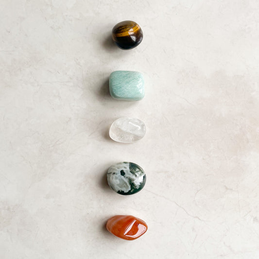 An arrangement of five tumbled crystals on a light surface, from top to bottom: black with hints of orange, translucent white, green with white patterns, clear with inclusions, and orange.