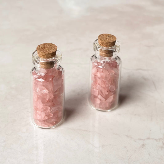 Two clear mini crystal jars with pink rose quartz chips, each with a cork lid, placed on a marble surface